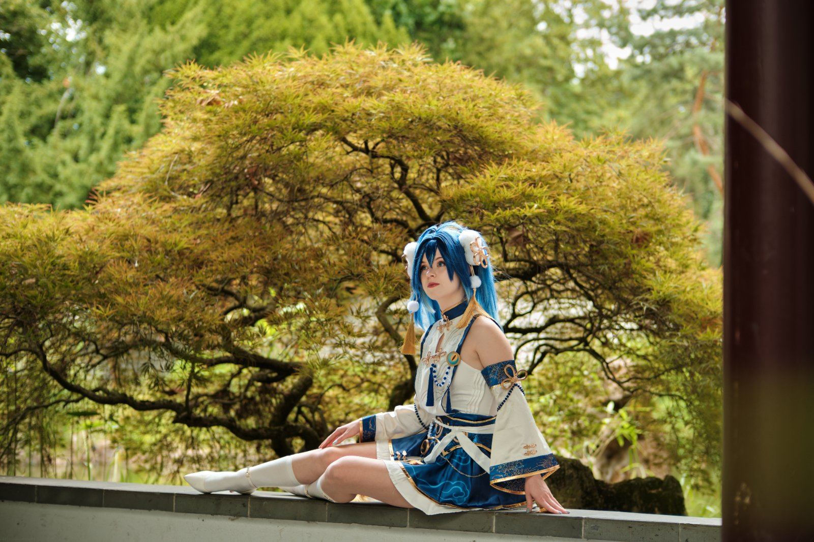 Cosplayerin mit blauer Perücke in blau-weißem Kostüm, sitzend vor einem japanischen Garten