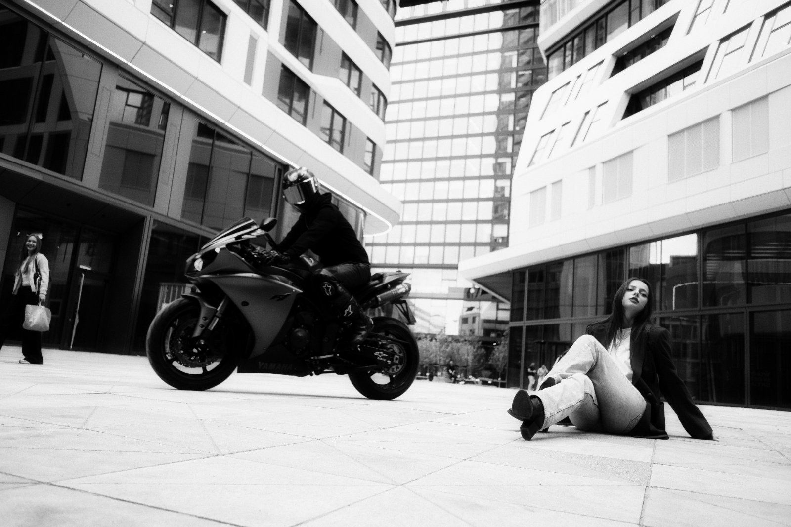 Schwarz-Weiß-Aufnahme: Frau sitzt entspannt auf dem Boden zwischen Hochhäusern, im Hintergrund fährt ein Motorrad vorbei