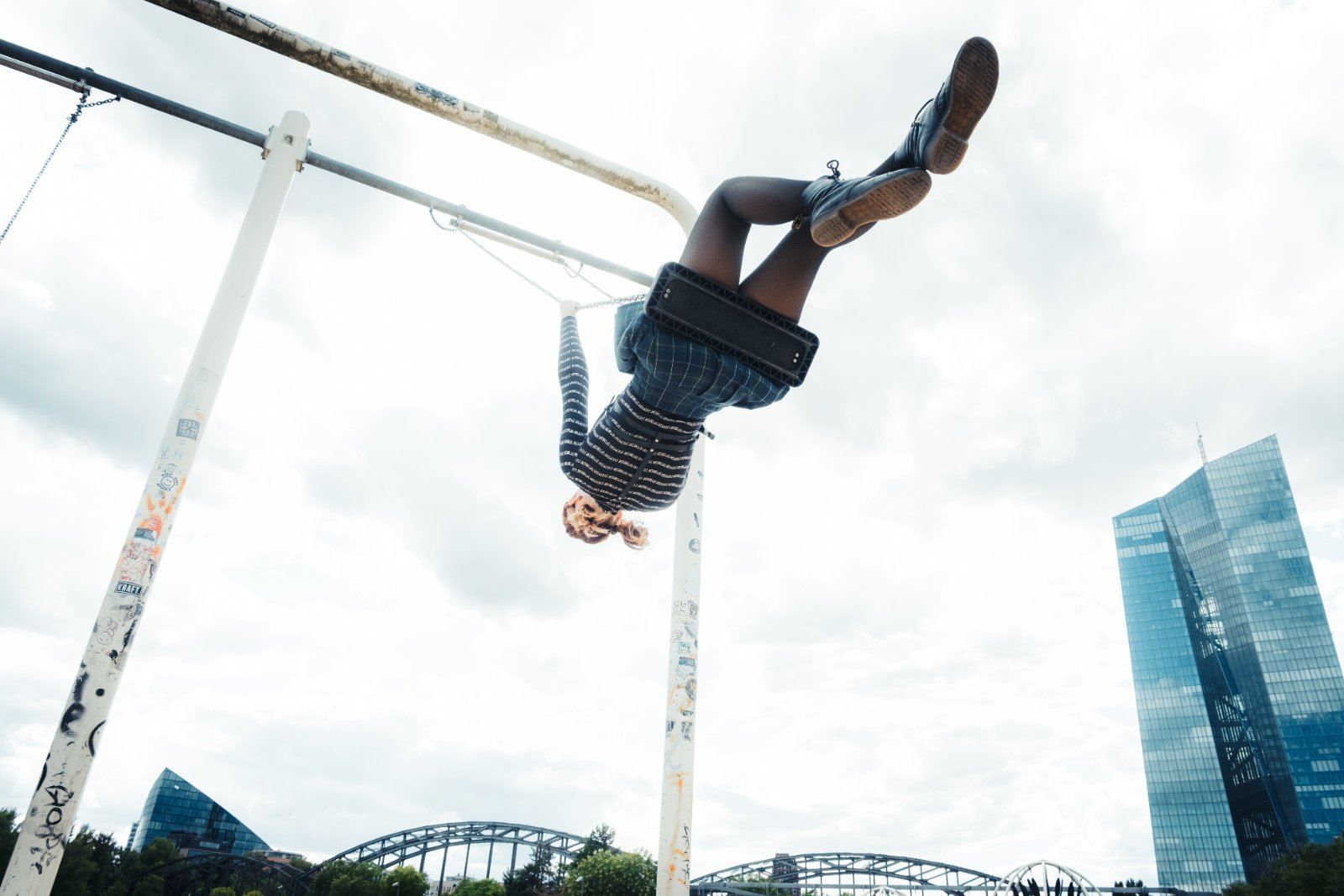 Person hängt kopfüber lachend an einer Parkschaukel vor der Frankfurter Skyline